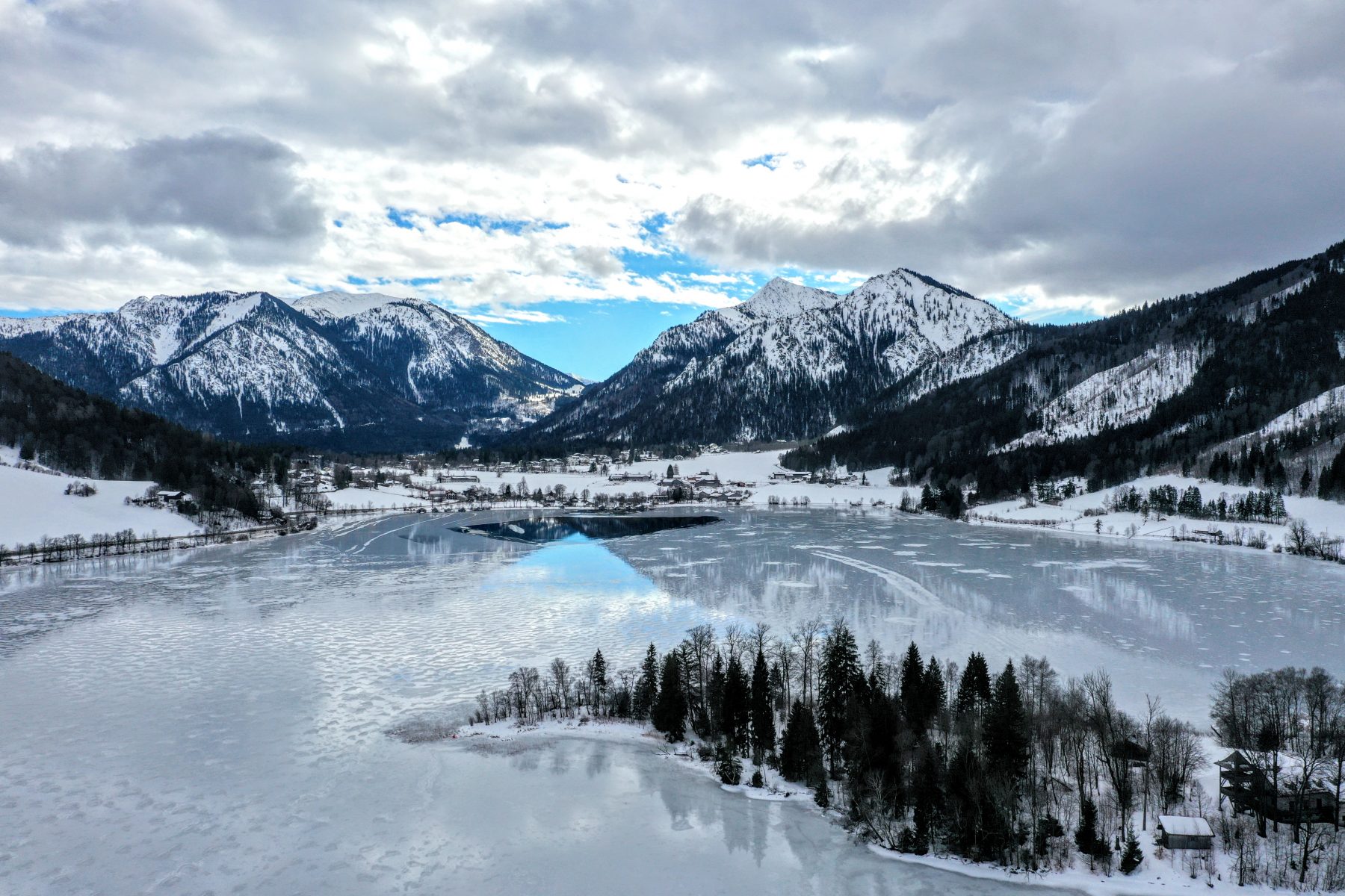 Blick auf den Schliersee im Winter, außenrum liegt Schnee, der Himmel spiegelt sich im Wasser.
