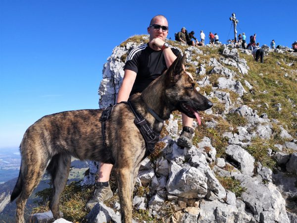 Ein Mann und sein Schäferhund sitzen auf einem Berg unter dem Gipfelkreuz.