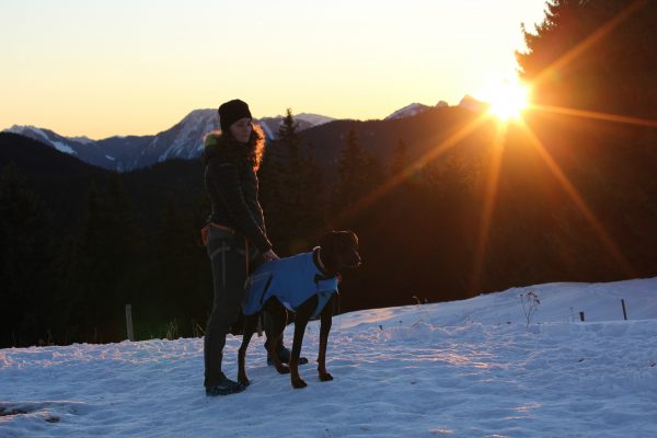Frau mit Hund stehen zum Sonnenuntergang im Schnee in den Bergen