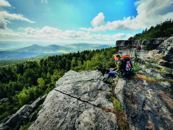 Eine Frau mit großem Rucksack sitzt hoch oben auf einem Felsen. Daneben ihr Hund