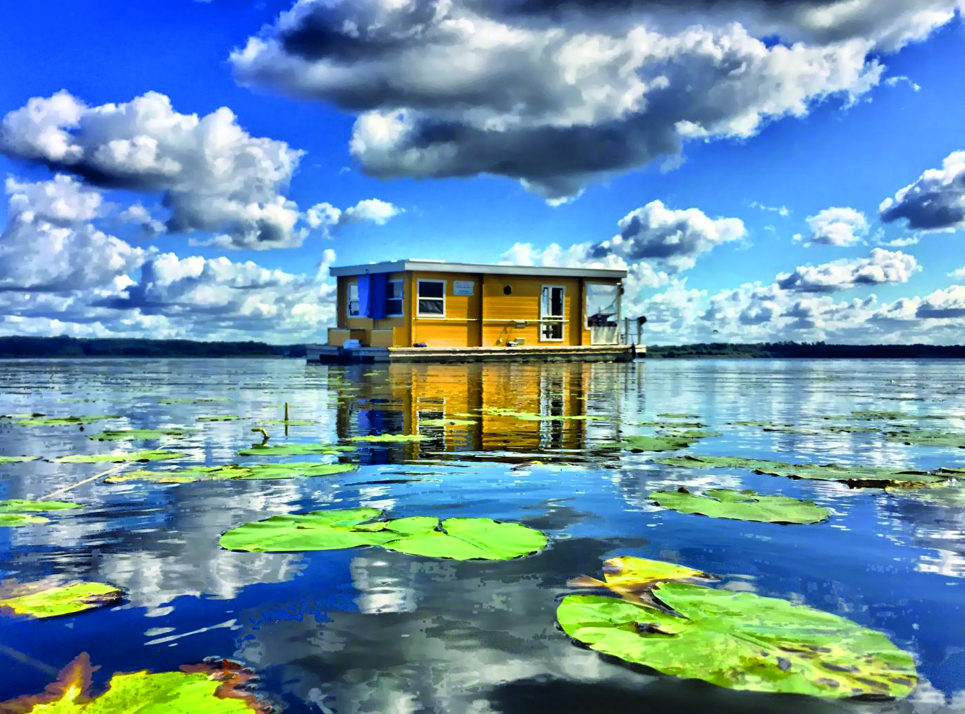 Ein gelbes Hausboot, umgeben von Seerosen im strahlendem Sonnenschein.