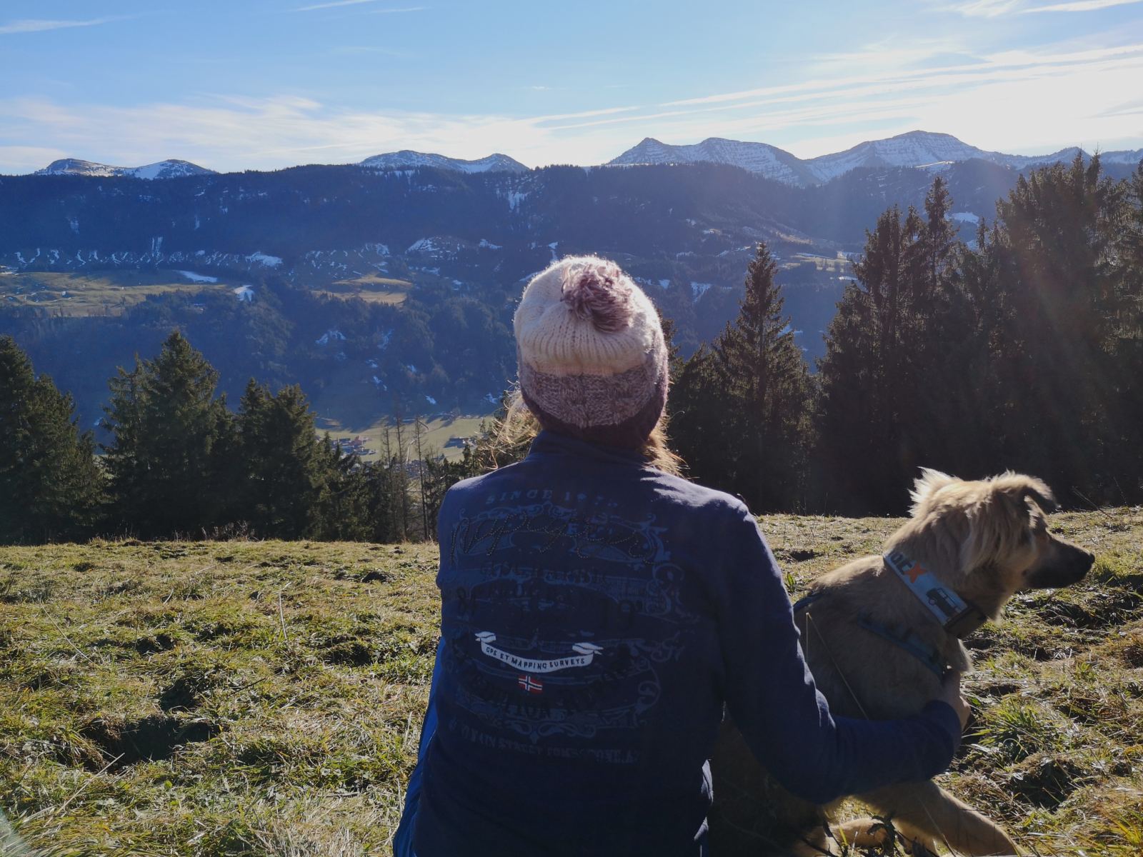 Eine Frau mit Mütze und einem kleinen Hund macht eine Pause auf einer Herbstwanderung. Sie blicken Richtung Berge.