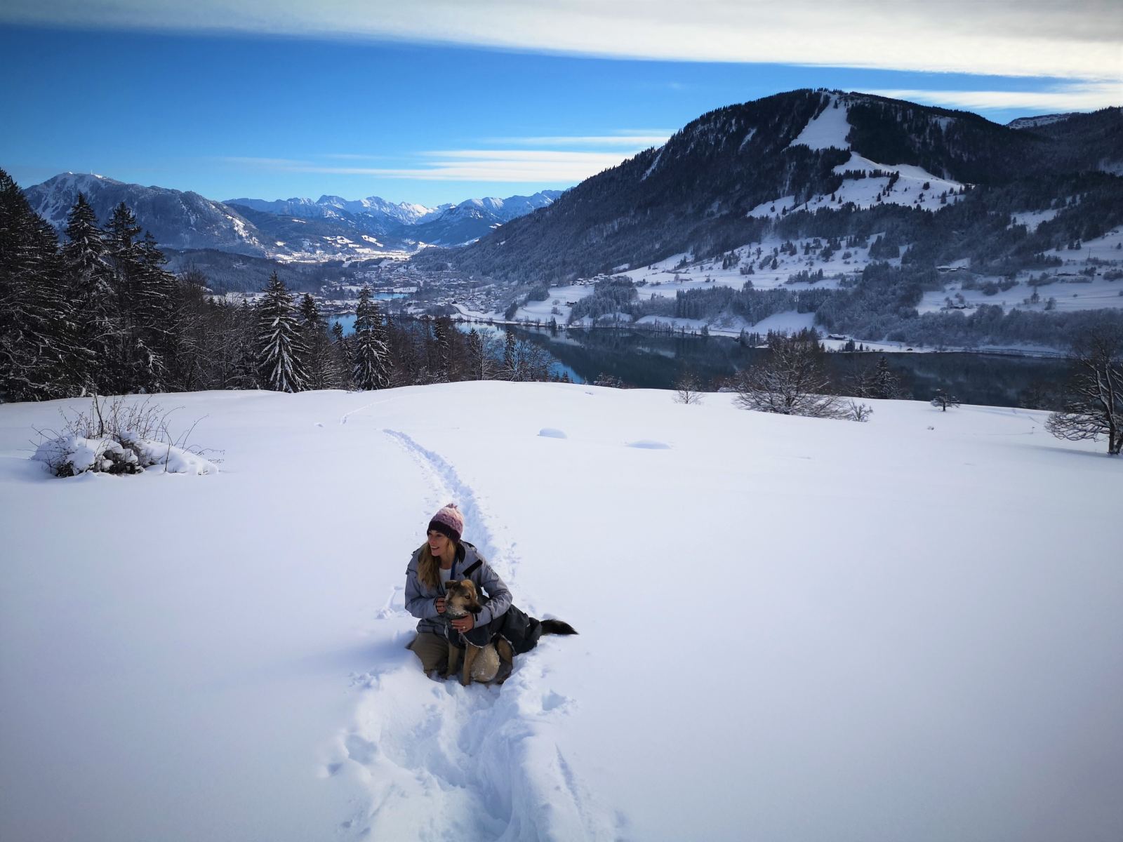 Eine Frau mit Pudelmütze knuddelt ihren Hund. Im Hintergrund die Berge, der Schnee abseits des Trampelpfades liegt hoch und unberührt.