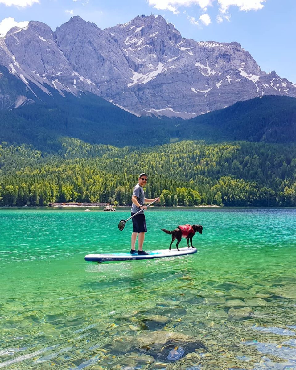 Ein Mann auf seinem SUP vor traumhafter Bergkulisse. Vorne auf dem Board steht ein kleiner schwarzer Hund mit Schwimmweste,